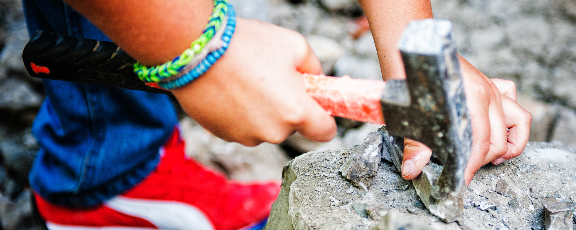 Zwei Kinderhände mit einem Hammer in der Hand, mit dem auf einen Stein geklopft wird.