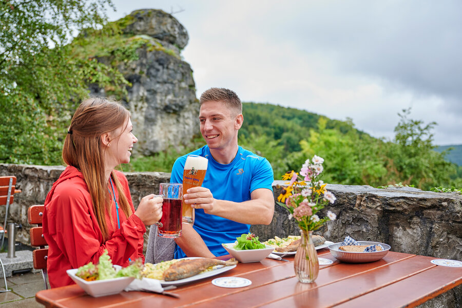 Eine junge Frau und ein junger Mann prosten sich an einem Tisch im Freien mit den Biergläsern zu und lächeln.