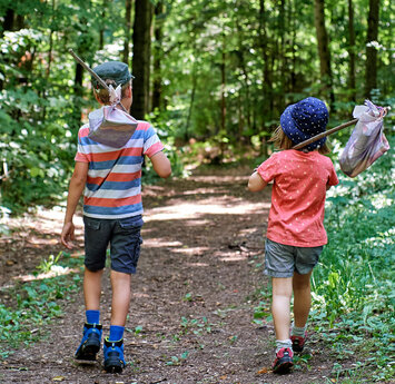Ein junge mit gestreiften T-Shirt und ein Mädchen mit rotem T-Shirt laufen auf einem Wanderweg durch den Wald. Auf den Schultern haben beide einen Stock, an dessen Ende ein kleiner Beutel hängt.