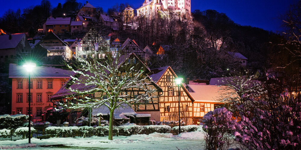 Im Vordergrund eine verschneite Parkanlage mit Beleuchtung im Winter. Im Hintergrund der Ort, über dem die beleuchtete Burg thront.