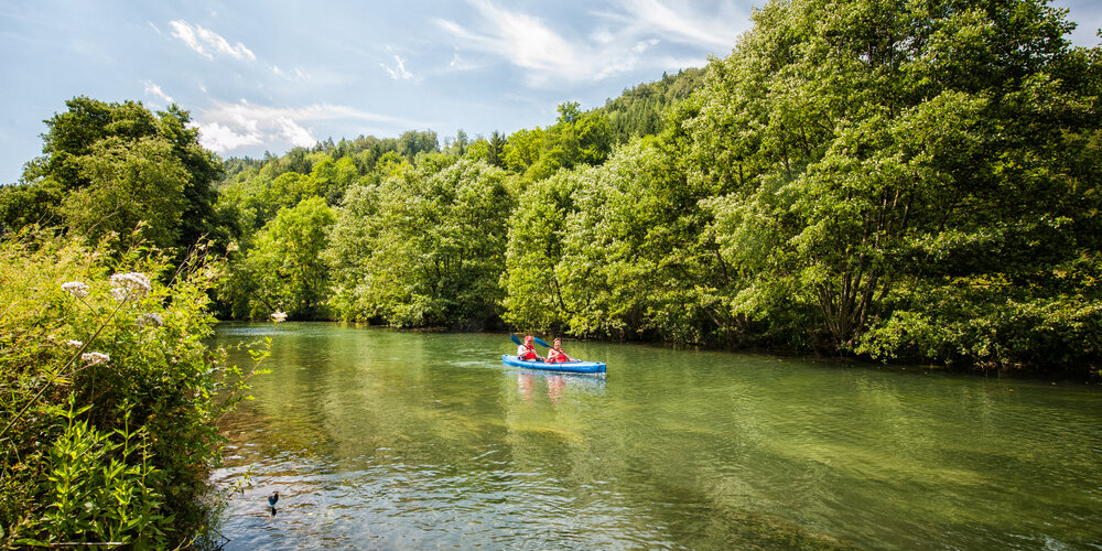 Ein Mann und eine Frau paddeln in einem blauen Kajak auf einem ruhigen Fluss.