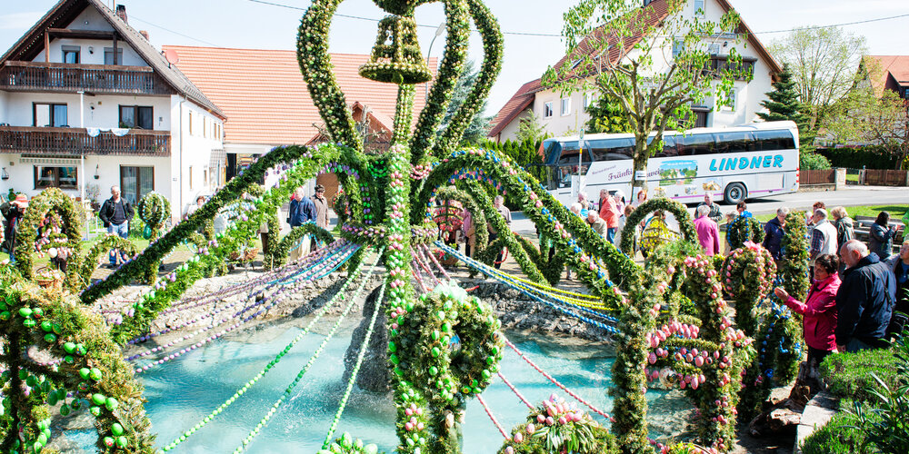 Ein bunt geschmückter Osterbrunnen mit vielen Ostereiern und grünen Girlanden wird von Besuchern bewundert.