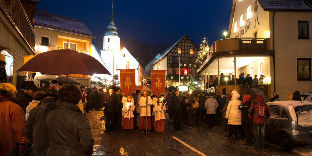 Eine Prozession, angeführt von mit Ministranten, die Fahnen tragen, laufen auf einer vereisten Straße vor einer Kirche. Am Straßenrand stehen viele Besucher.