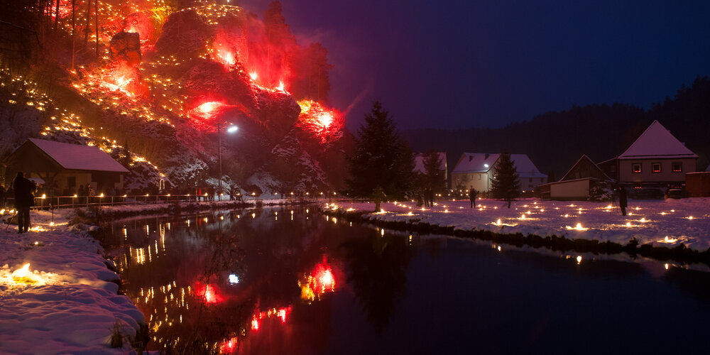 Ein ruhiger Bach fließt durch den Talgrund. Auf den angrenzenden Wiesen werden kleine Feuer entfacht und Menschen stehen daneben. Im Hintergrund erhebt sich ein Felshang mit vielen roten Feuern und einem leuchteten Kreuz.