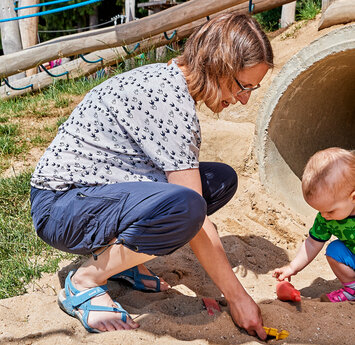 Ein Kind sitzt an einem Spielplatz vor einer Röhre und spielt im Sand. Die Mutter kniet neben dem Kind.