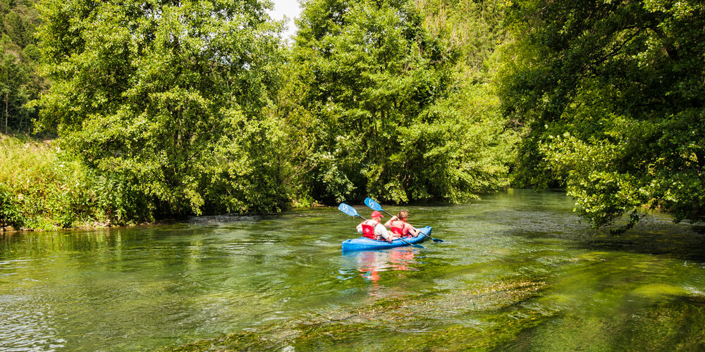 Auf einem Fluss fahren zwei Kanuten in einem blauen Kanu. Am Flussufer stehen Bäume.