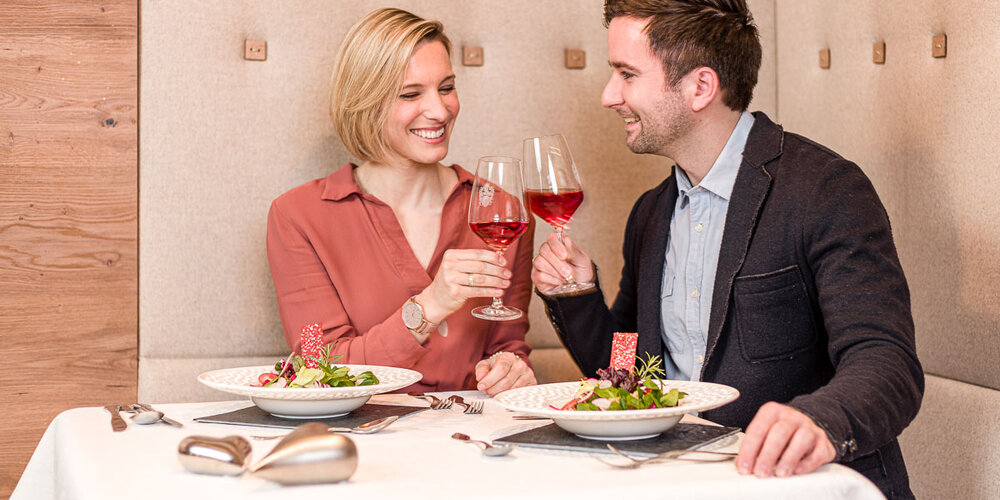 Ein Paar sitzt lächelnd an einem Tisch und prostet sich mit den Weingläsern zu. Auf dem Tisch stehen zwei Teller mit Salat.