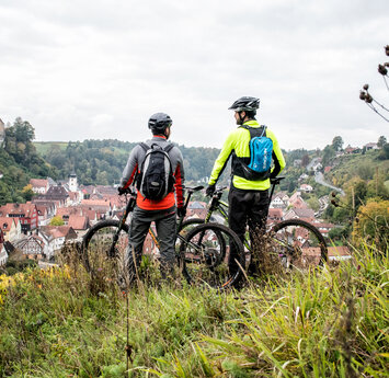 Zwei Männer von hinten stehen mit ihren Mountainbikes über dem Ort und blicken hinab.