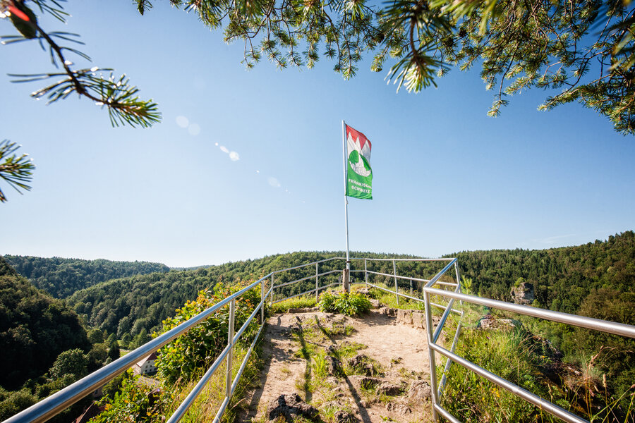 Ein schmaler, felsiger Weg, der von einem Geländer umgeben ist, führt zu einem Aussichtspunkt. Dort steht eine wehende Fahne.