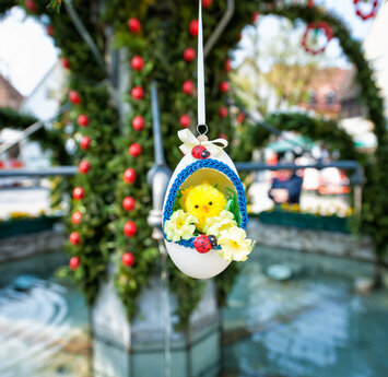 In einem aufgeschnittenen Ei sitzt ein kleines Kunststoffküken. Das Ei hängt an einer Schnur am Osterbrunnen, der im Hintergrund zu erahnen ist.