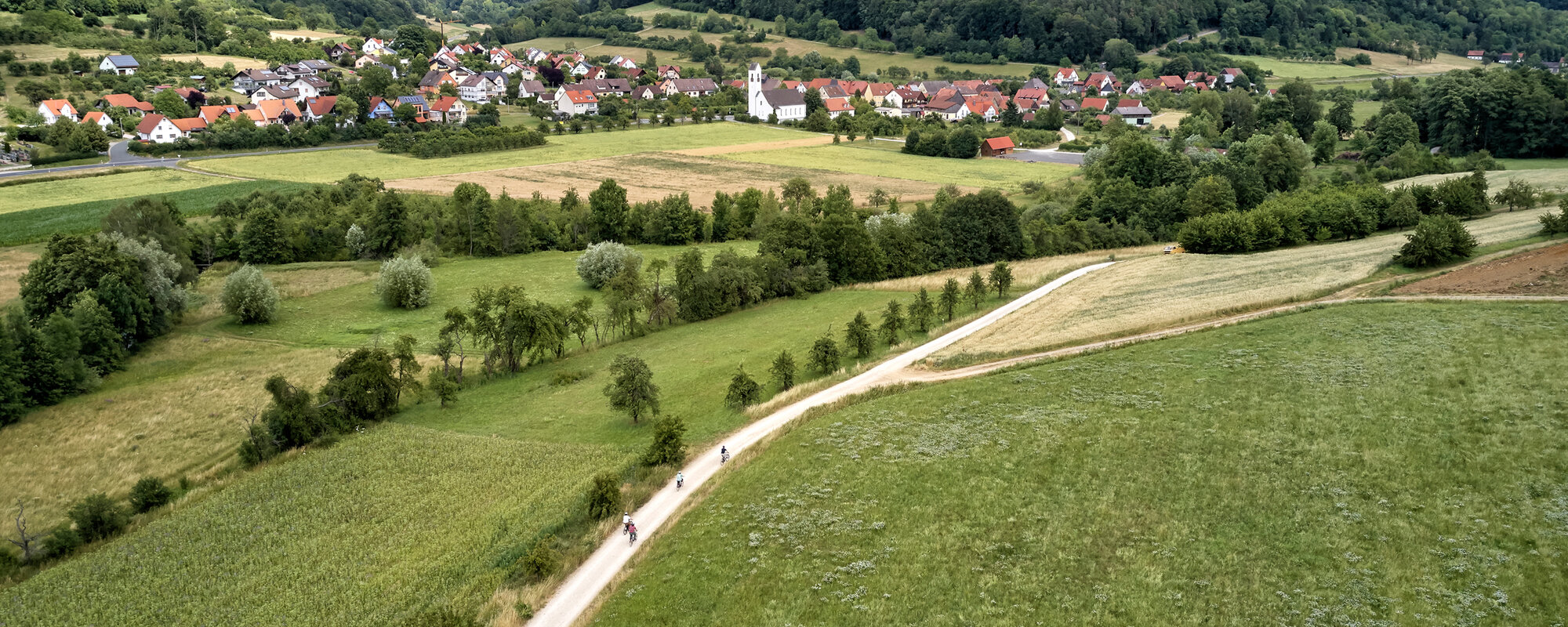 Im Vordergrund fahren vier Radfahrer auf einem Feldweg. Im Hintergrund ist ein Ort in Tallage vor bewaldeten Hügeln zu sehen.