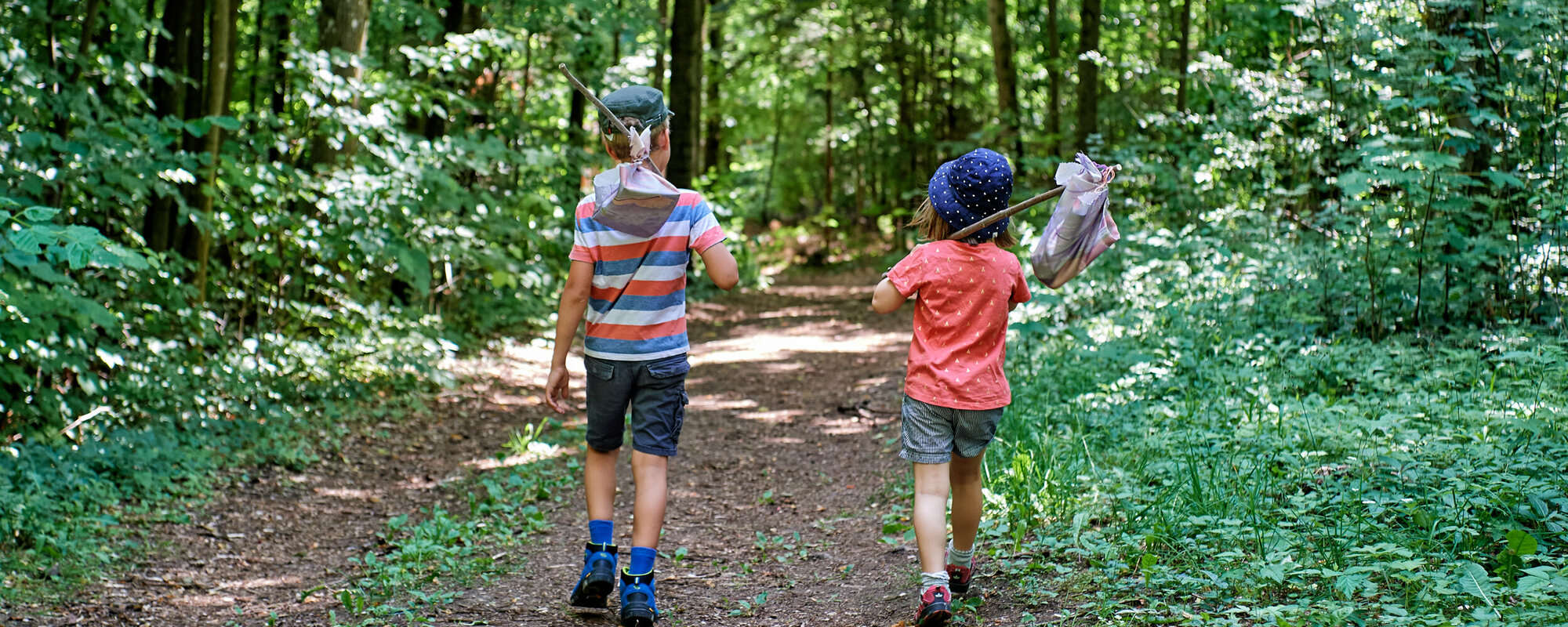 Ein junge mit gestreiften T-Shirt und ein Mädchen mit rotem T-Shirt laufen auf einem Wanderweg durch den Wald. Auf den Schultern haben beide einen Stock, an dessen Ende ein kleiner Beutel hängt.