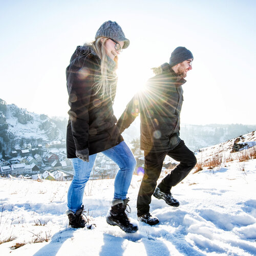 Ein lächelndes Paar läuft händchenhaltend oberhalb der Stadt durch den weißen Schnee.