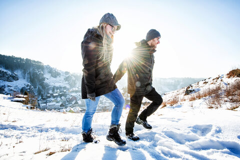 Ein lächelndes Paar läuft händchenhaltend oberhalb der Stadt durch den weißen Schnee.