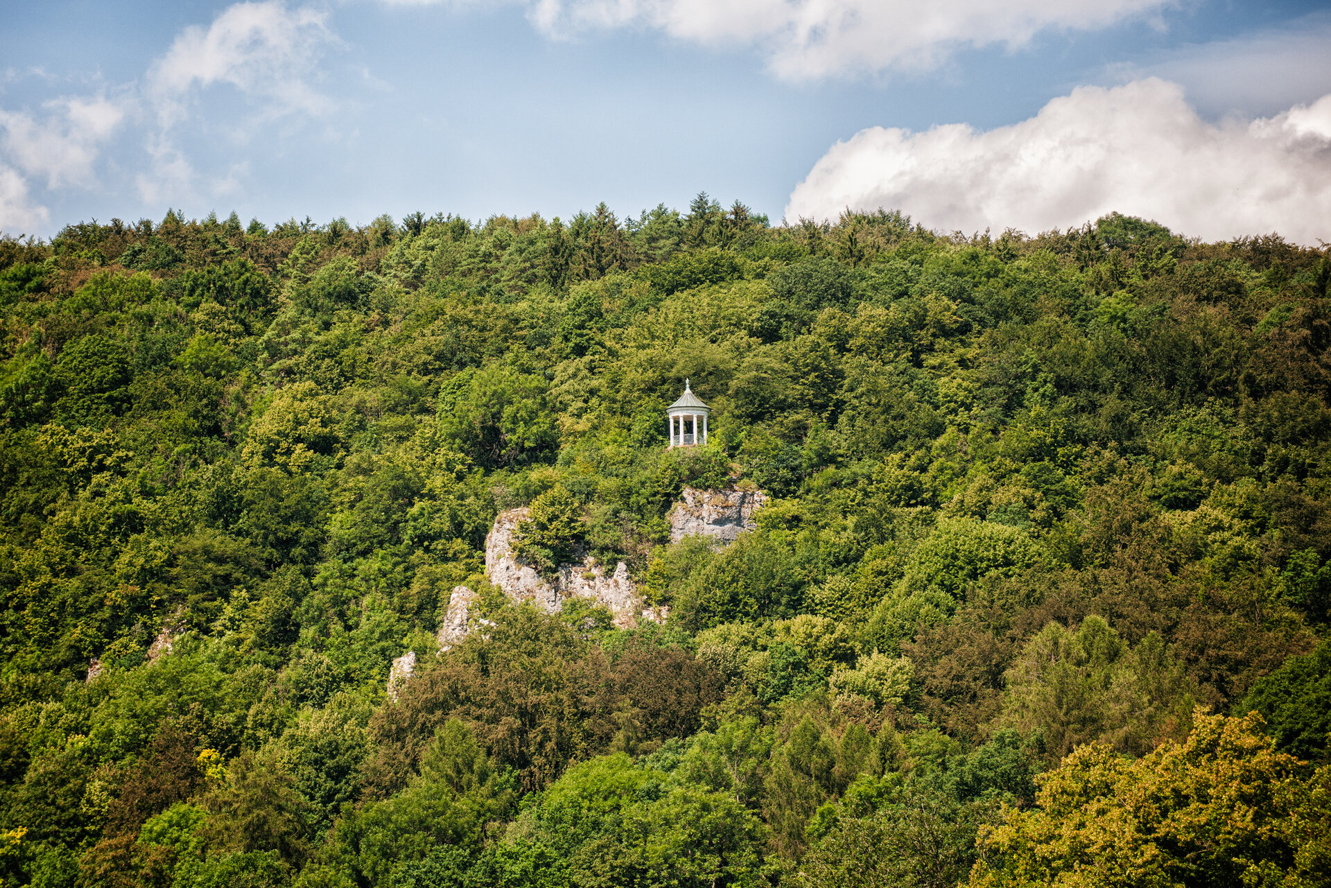 Ein bewaldeter Berg, aus dem graue Felsnadeln spitzen. Auf einer der Felsnadeln steht ein weißer, steinerner Aussichtspavillon.