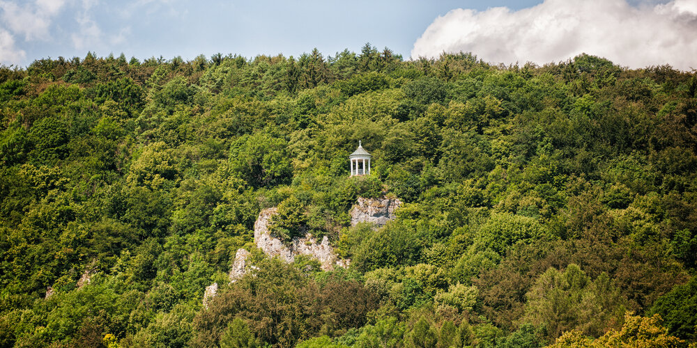 Ein bewaldeter Berg, aus dem graue Felsnadeln spitzen. Auf einer der Felsnadeln steht ein weißer, steinerner Aussichtspavillon.