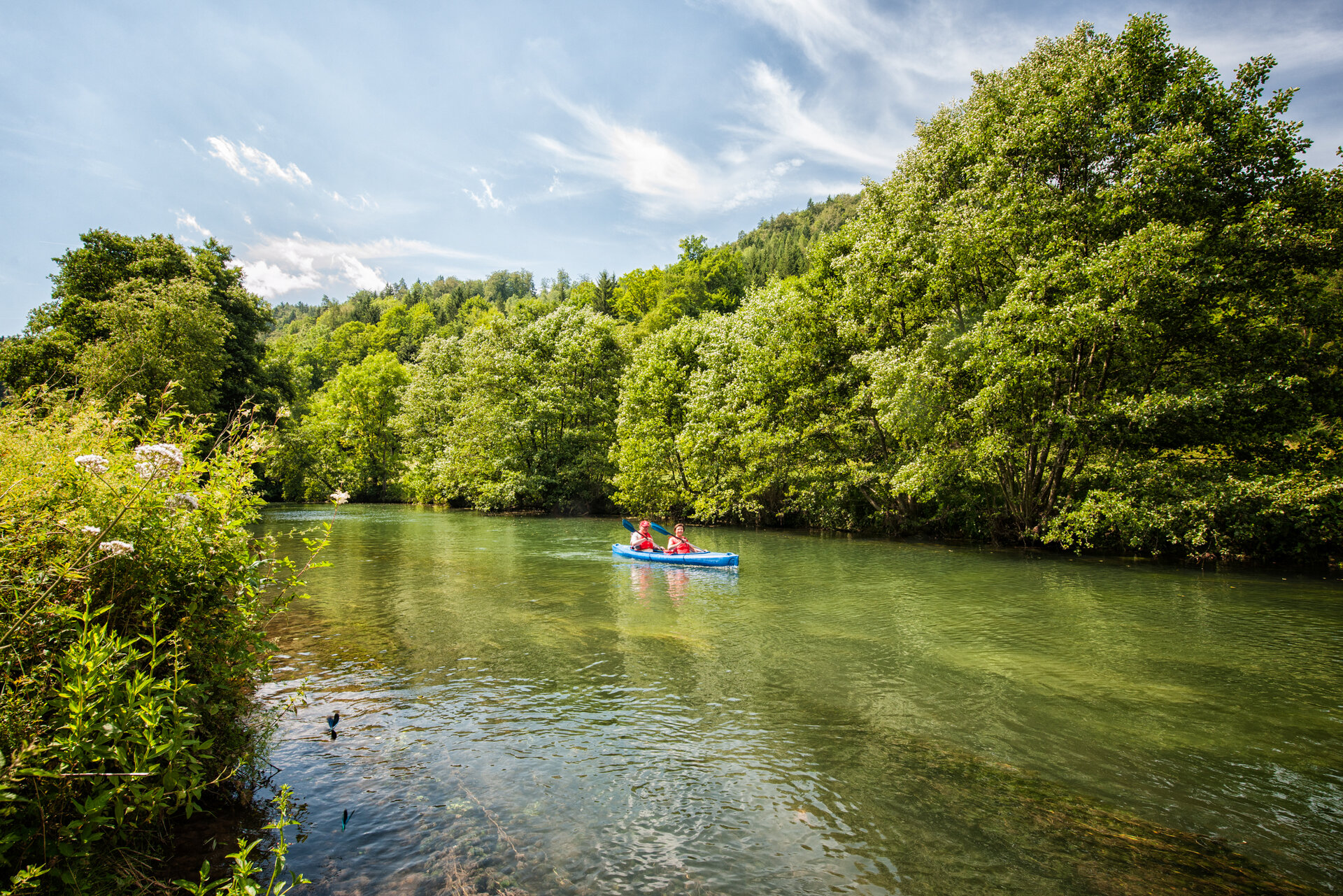 Ein Mann und eine Frau paddeln in einem blauen Kajak auf einem ruhigen Fluss.