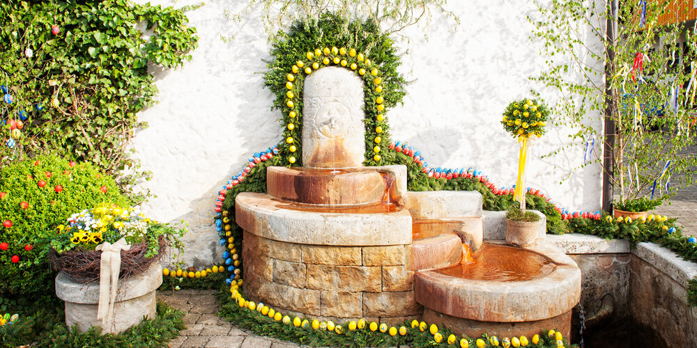 Ein fünfstufiger Brunnen mit kupferfarbenen Becken ist mit gelben, roten und blauen Ostereiern und Nadelzweigen geschmückt.