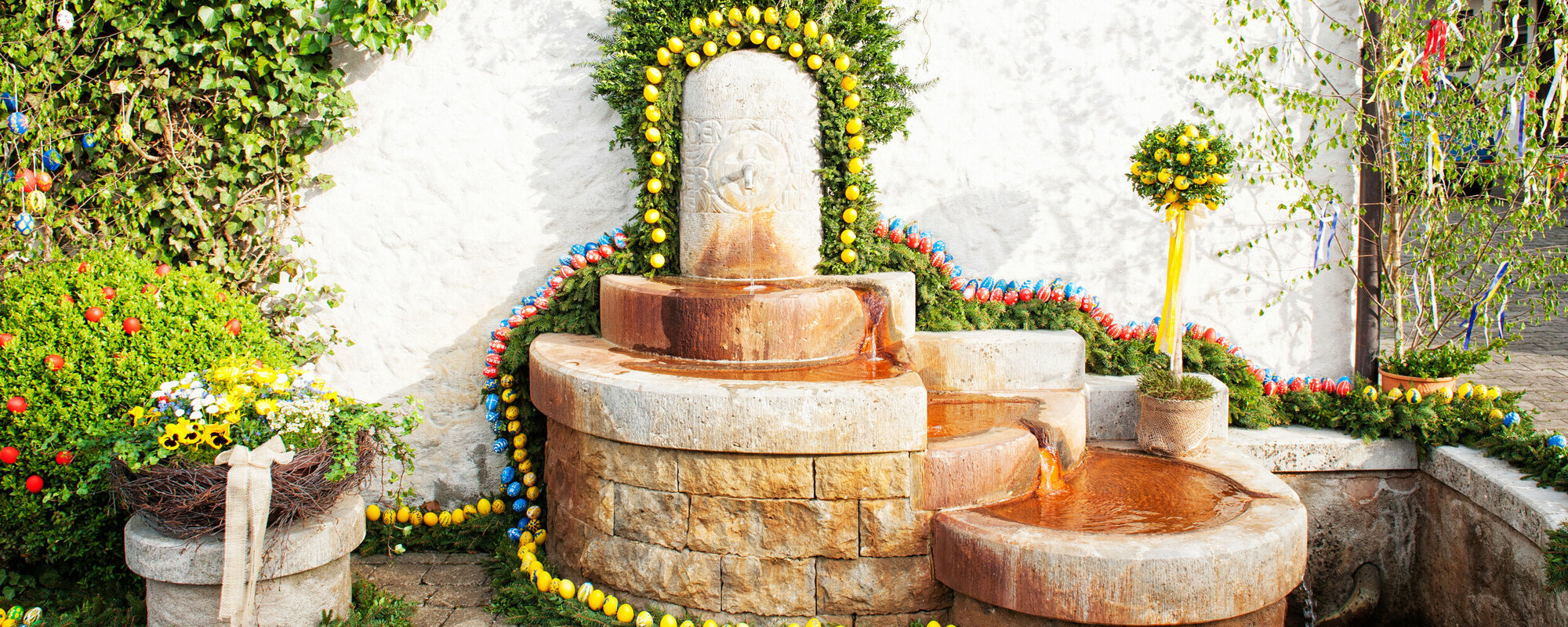 Ein fünfstufiger Brunnen mit kupferfarbenen Becken ist mit gelben, roten und blauen Ostereiern und Nadelzweigen geschmückt.