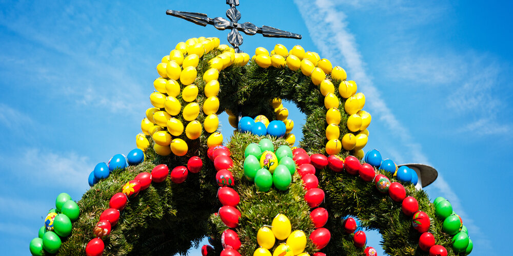 Mit bunten Eiern geschmückte Krone eines Osterbrunnens aus Grünzeug. Darauf erhebt sich ein Kreuz vor blauem Himmel.