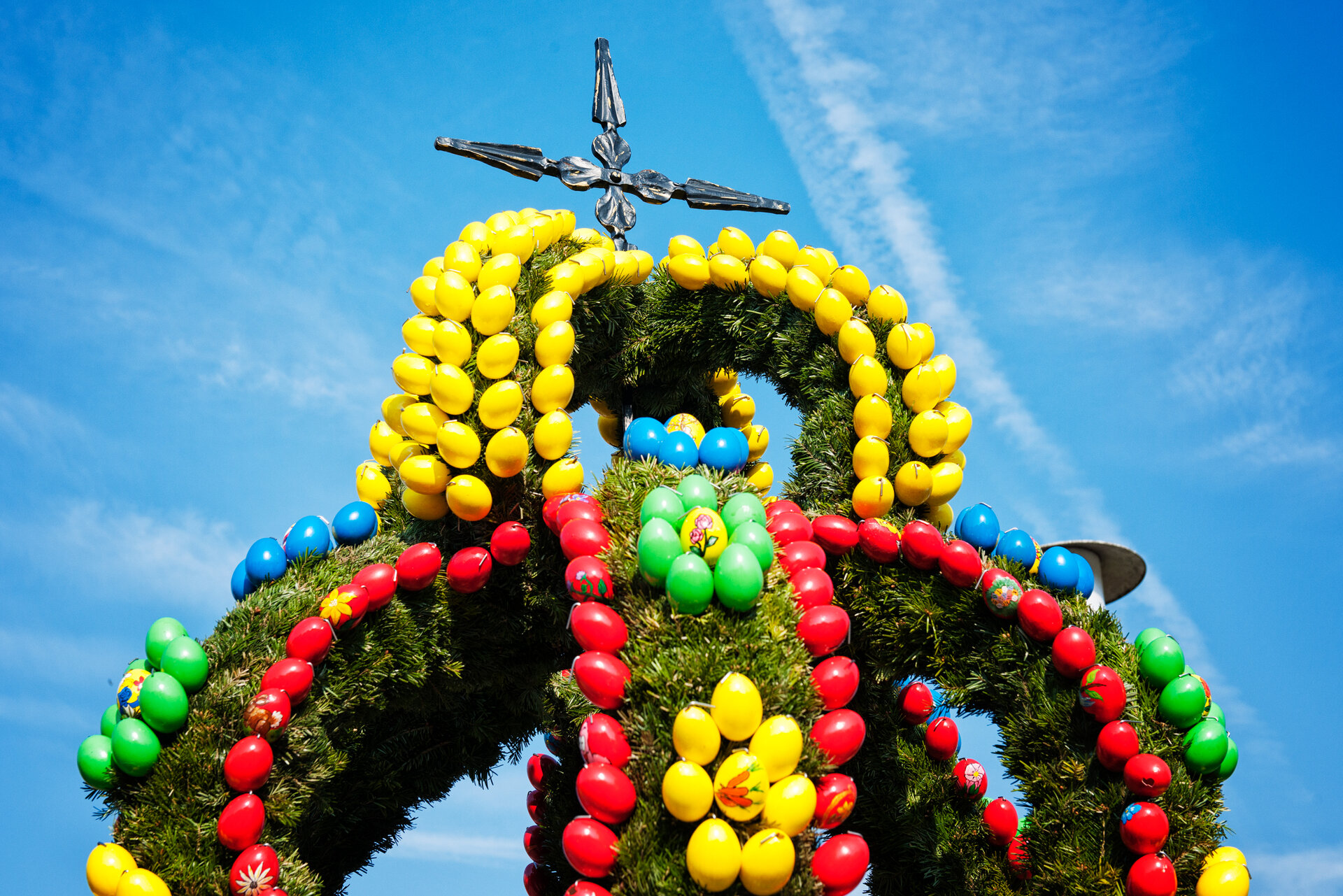 Mit bunten Eiern geschmückte Krone eines Osterbrunnens aus Grünzeug. Darauf erhebt sich ein Kreuz vor blauem Himmel.