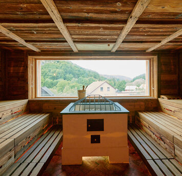 Eine Sauna mit Aufgussofen und Holzbänken auf der linken und rechten Seite. Ein Panoramafest ermöglicht die Sicht nach außen.