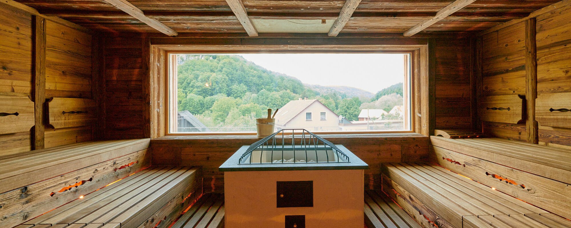 Eine Sauna mit Aufgussofen und Holzbänken auf der linken und rechten Seite. Ein Panoramafest ermöglicht die Sicht nach außen.