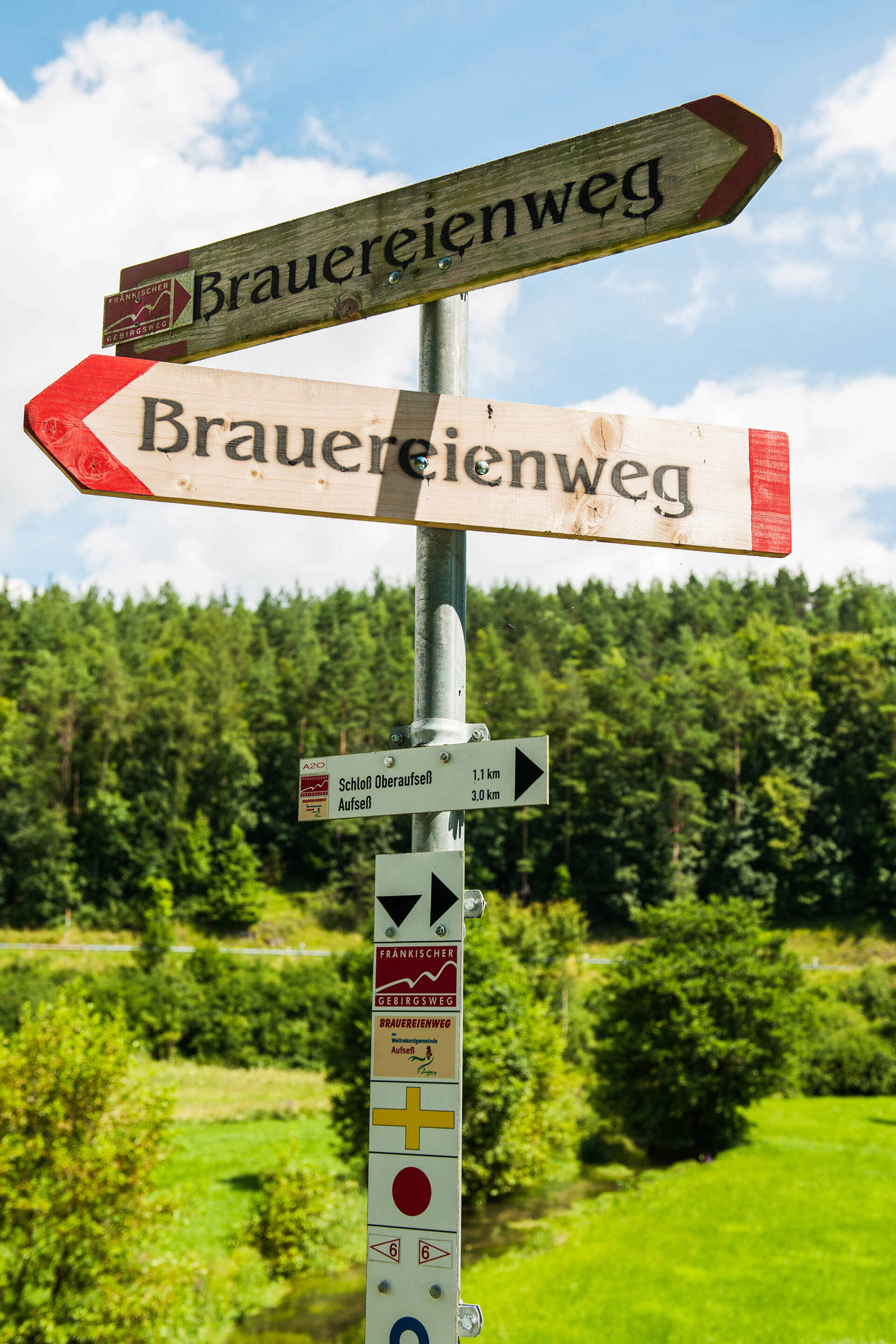 An einem Pfosten sind mehrer Schilder mit Wanderzeichen angebracht. Zwei große Hinweisschilder aus Holz tragen die Aufschrift Brauereienweg.