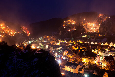 Blick hinab auf den Talkessel von Pottenstein bei Nacht. Die Holzfeuer an den Berghängen brennen. Die Straßen im Ort sind erleuchtet.