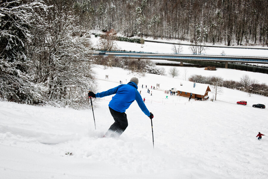 Ein Wintersportler mit blauer Jacke fährt auf Skiern den Berg hinab. Im Hintergrund ist das Lifthäuschen zu sehen.