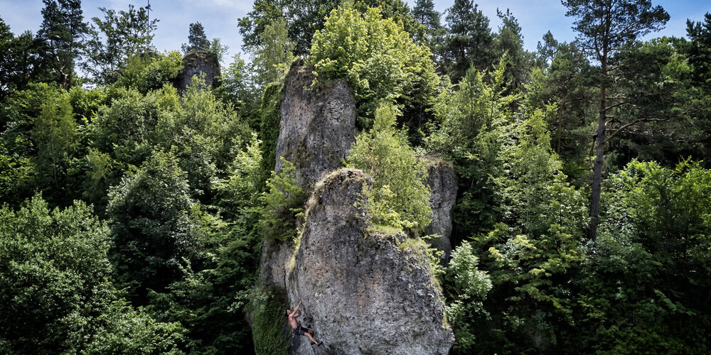 An einem grauen Felsen klettert ein Mann mit freiem Oberkörper. Der Felsen wird von grünem Wald umfasst.