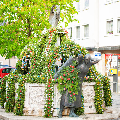 Ein sechseckiger Brunnen mit Bronzefigur. Über den Brunnen hängen grüne Girlanden mit bemalten Ostereiern. 