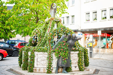 Ein sechseckiger Brunnen mit Bronzefigur. Über den Brunnen hängen grüne Girlanden mit bemalten Ostereiern. 