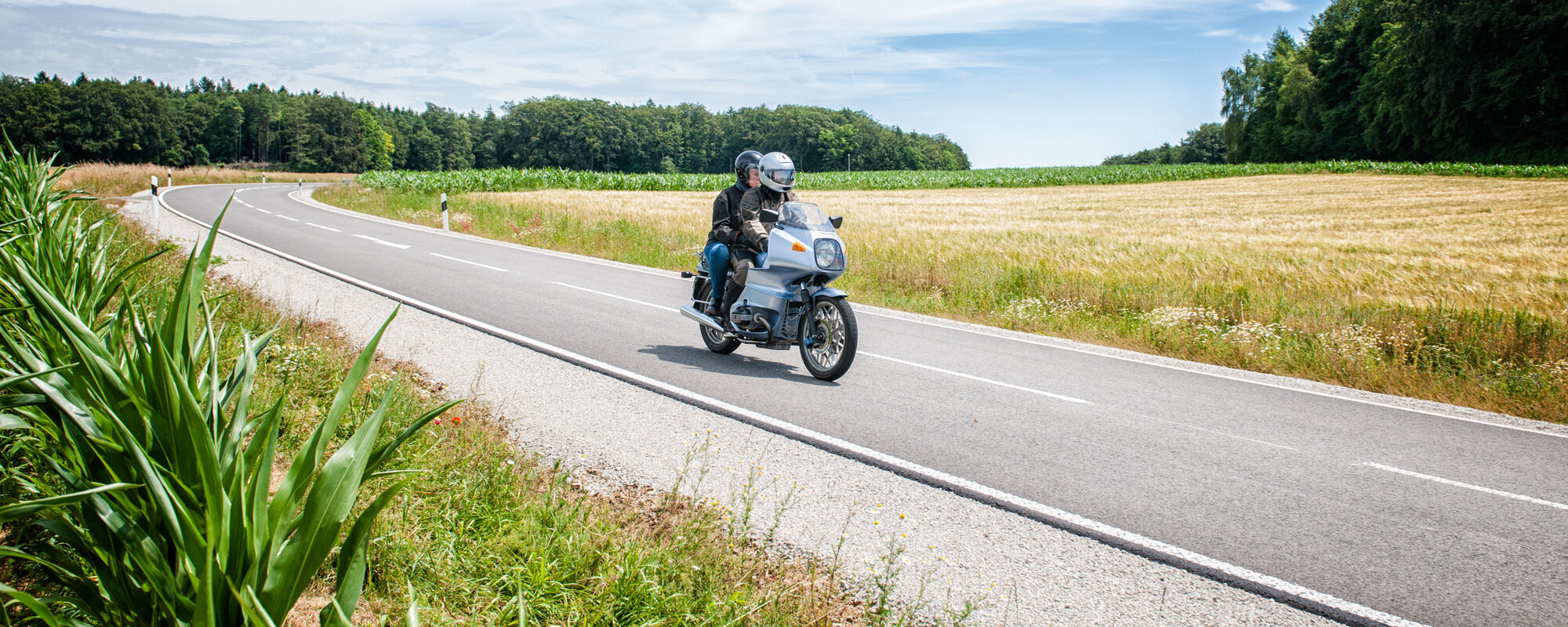 Zwei Personen auf einem Motorrad fahren auf einer Straße zwischen Wiesenfeldern.