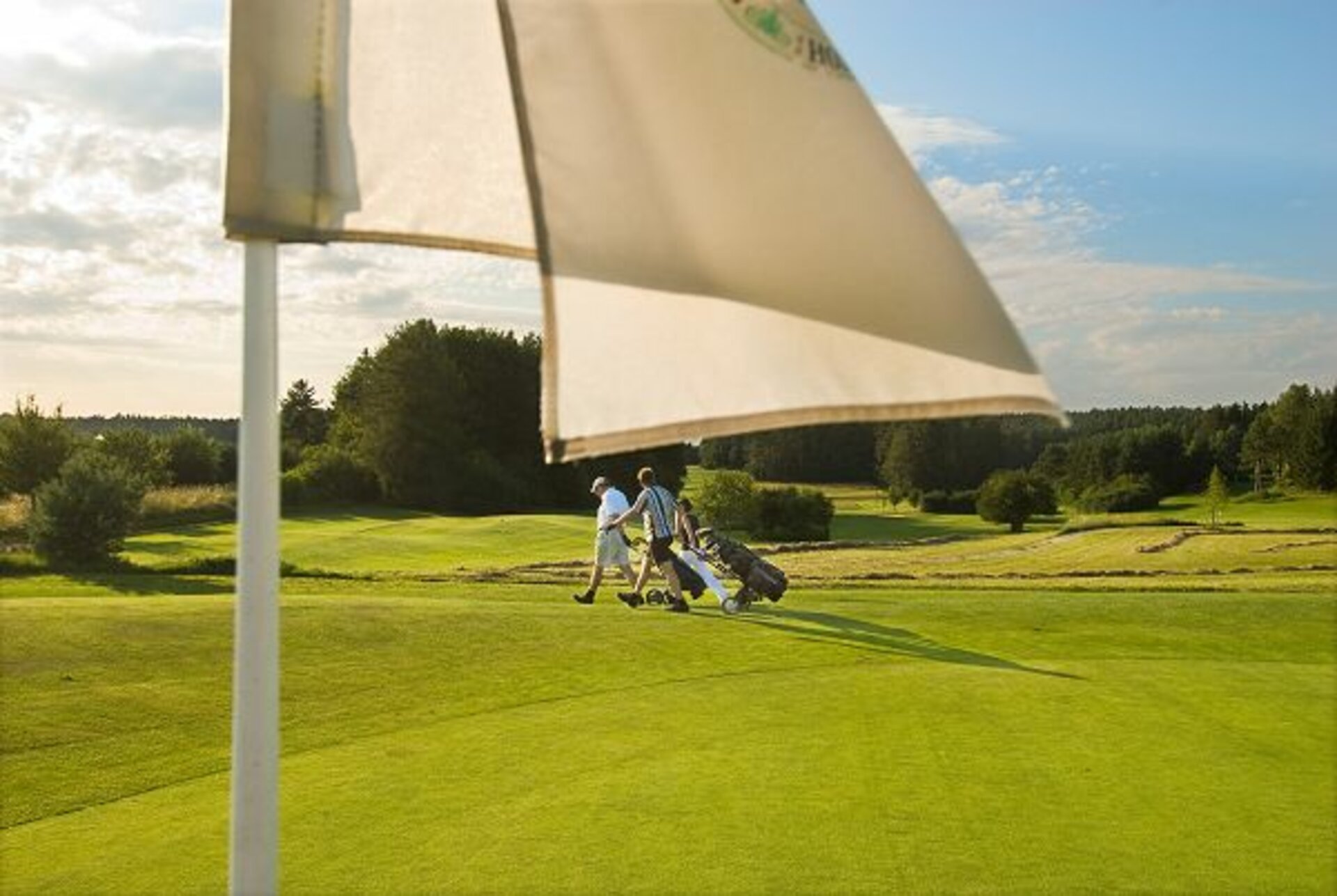 Vor dem saftigen Grün eines Golfplatzes steht ein weißes Fähnchen. Im Hintergrund laufen zwei Golfspieler mit dem Wagen über den Platz.
