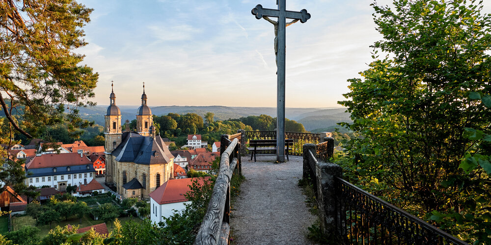 Ein Schotterweg, der auf beiden Seiten von einem Geländer eingefasst wird, führt zu einem Aussichtspunkt mit Kreuz. Im Hintergrund ist die große, barocke Kirche zu sehen.