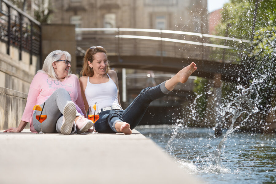 Zwei Frauen sitzen an einer künstlichen Uferpromenade und lachen. Eine davon spritzt mit dem Fuß das Wasser aus dem Bach in die Luft.