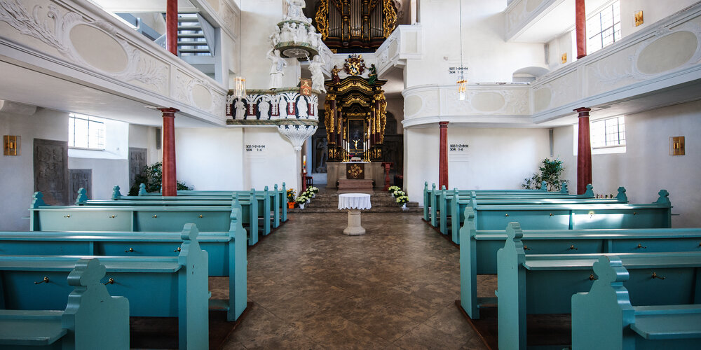 Blick auf den Altar der Kirche, vor dem türkise Kirchenbänke stehen. Links und rechts oberhalb sind jeweils zwei übereinanderliegende Emporen.