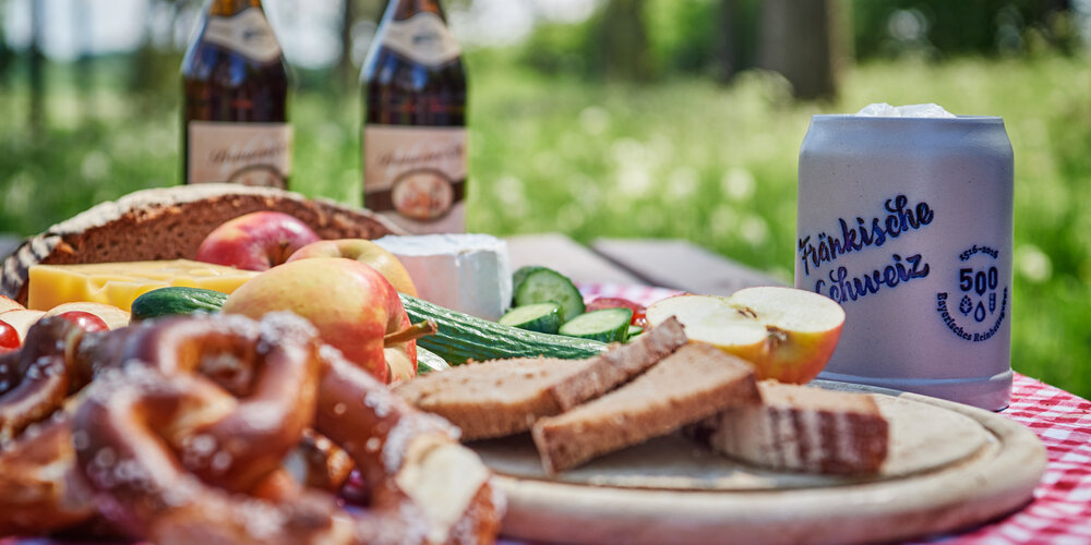 Detailaufnahme einer vegetarischen Brotzeit mit Brezl, Brot, Käse, Obst und Gemüse. Im Hintergrund stehen zwei Falschen Bier und ein Steinkrug.