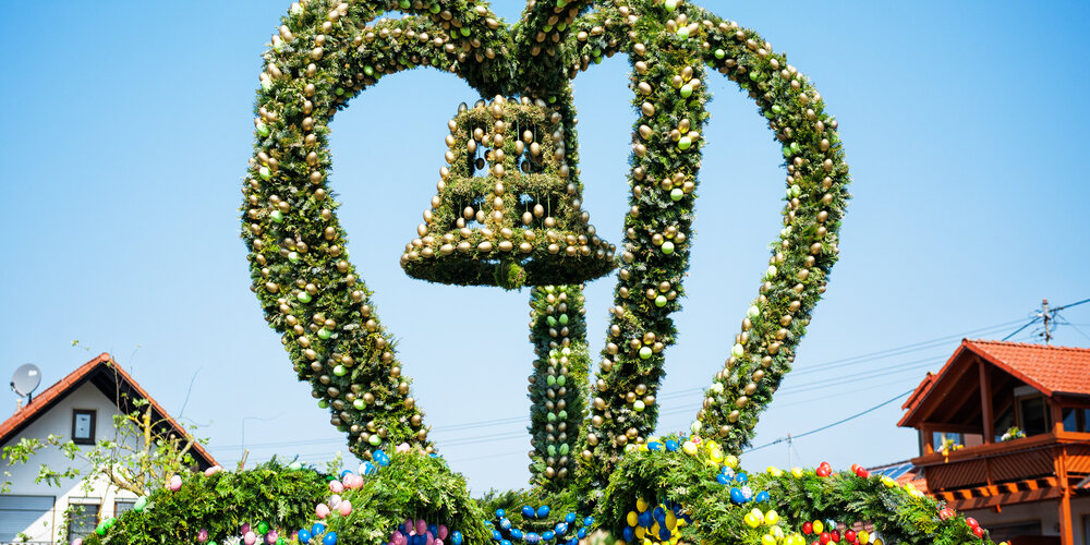 Eine aus grünen Zweigen gebastelte Krone auf dem Osterbrunnen. Sie ist mit goldenen Eiern geschmückt. In der Mitte der Krone hängt eine Glocke.