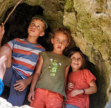 In einer Felswand ist ein großes Loch in dem zwei Mädchen und ein Junge mit einem Stock in der Hand stehen.