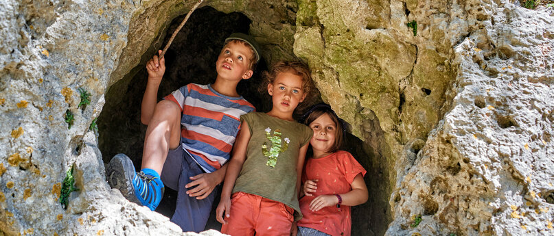 In einer Felswand ist ein großes Loch in dem zwei Mädchen und ein Junge mit einem Stock in der Hand stehen.
