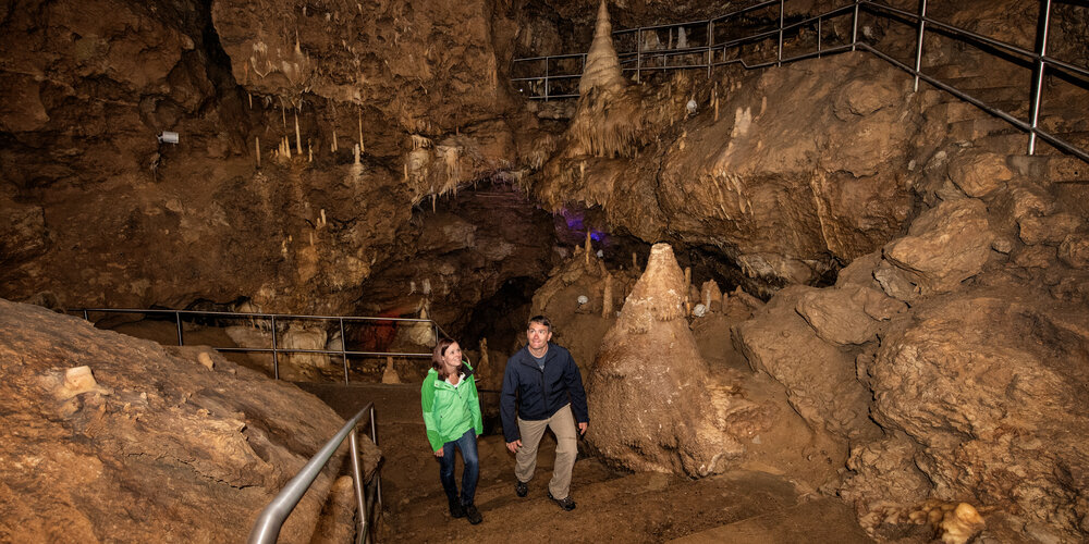 Ein Paar läuft lächelnd die Treppen in der Höhle hinauf. Sie befinden sich in einem großen Saal mit vielen Tropfsteinen und bewundern diese.