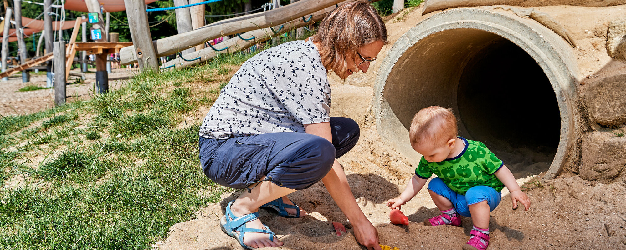 Ein Kind sitzt an einem Spielplatz vor einer Röhre und spielt im Sand. Die Mutter kniet neben dem Kind.
