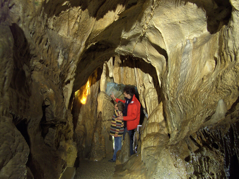 Ein Mann in roter Jacke und ein Kind stehen sehr nahe an einem Tropfstein in einer Höhle.