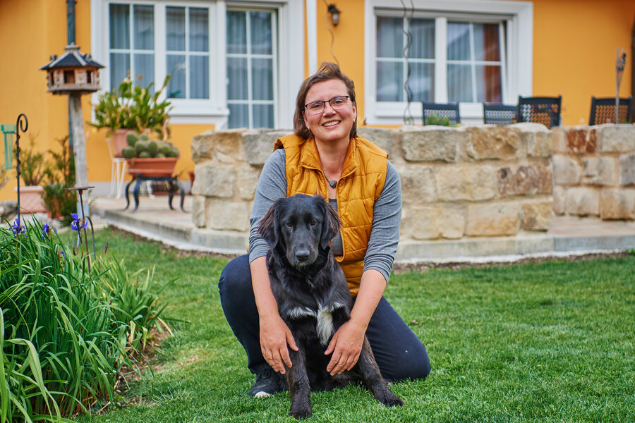 Eine Frau mit gelber Weste kniet mit ihrem schwarzen Hund in der Wiese vor einem Haus.