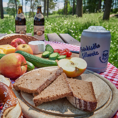 Auf einem Rasttisch im Wald ist ein vegaritscher Brotzeitteller mit Käse, Gemüse und Obst angerichtet. Dazu stehen zwei Flaschen Bier und ein Steinkrug im Hintergrund.