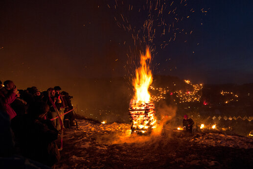 Eine große Gruppe Menschen steht im Duklen neben einem brennenden Holzfeuer. Im Hintergrund erleuchten die Lichter der Stadt.