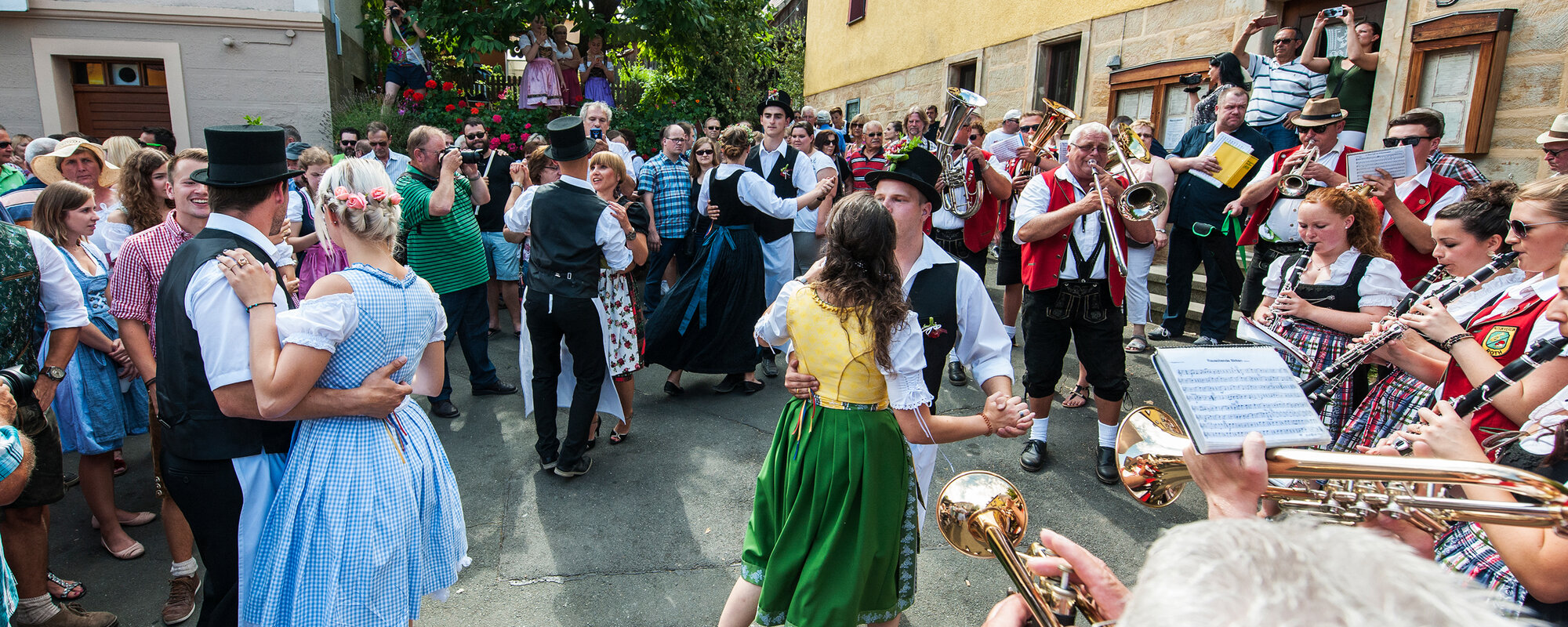 Am Dorfplatz feiert eine große Gruppe von Menschen, die teilweise fränkische Tracht tragen. Eine Musikkapelle mit roten Westen spielt.