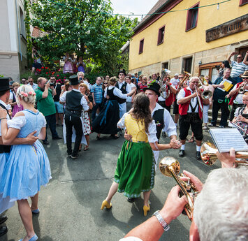 Am Dorfplatz feiert eine große Gruppe von Menschen, die teilweise fränkische Tracht tragen. Eine Musikkapelle mit roten Westen spielt.